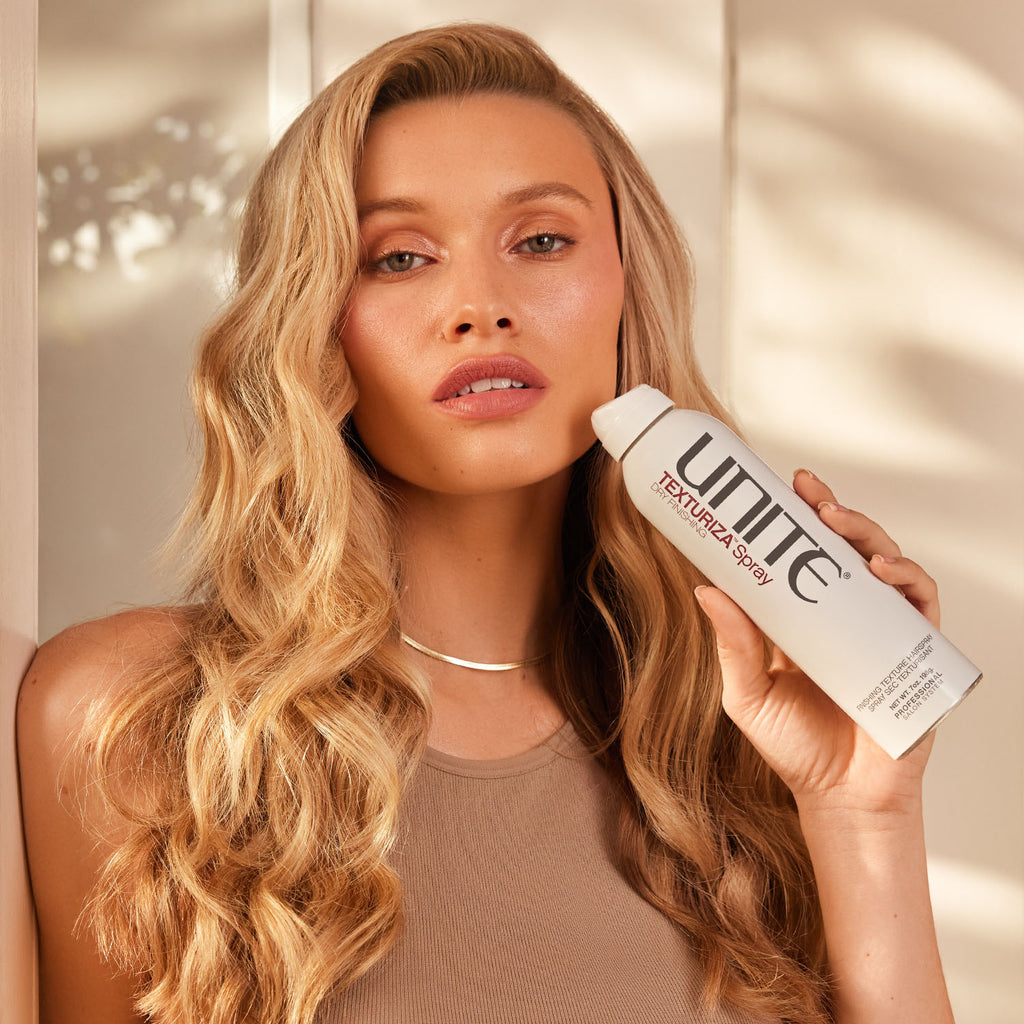 Blonde woman with wavy hair holding a bottle of TEXTURIZA Hair Texturizing Spray.