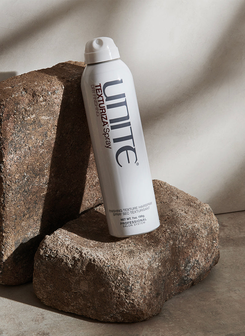 Bottle of texturizing spray hair product displayed on stone blocks in natural light.