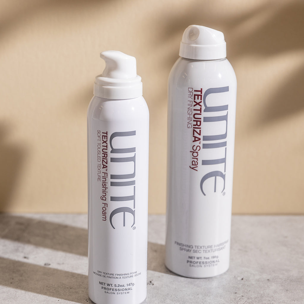 Two bottles of UNITE Hair’s products, one labeled TEXTURIZA Finishing Foam and the other TEXTURIZA Spray, were placed on a light-colored surface.